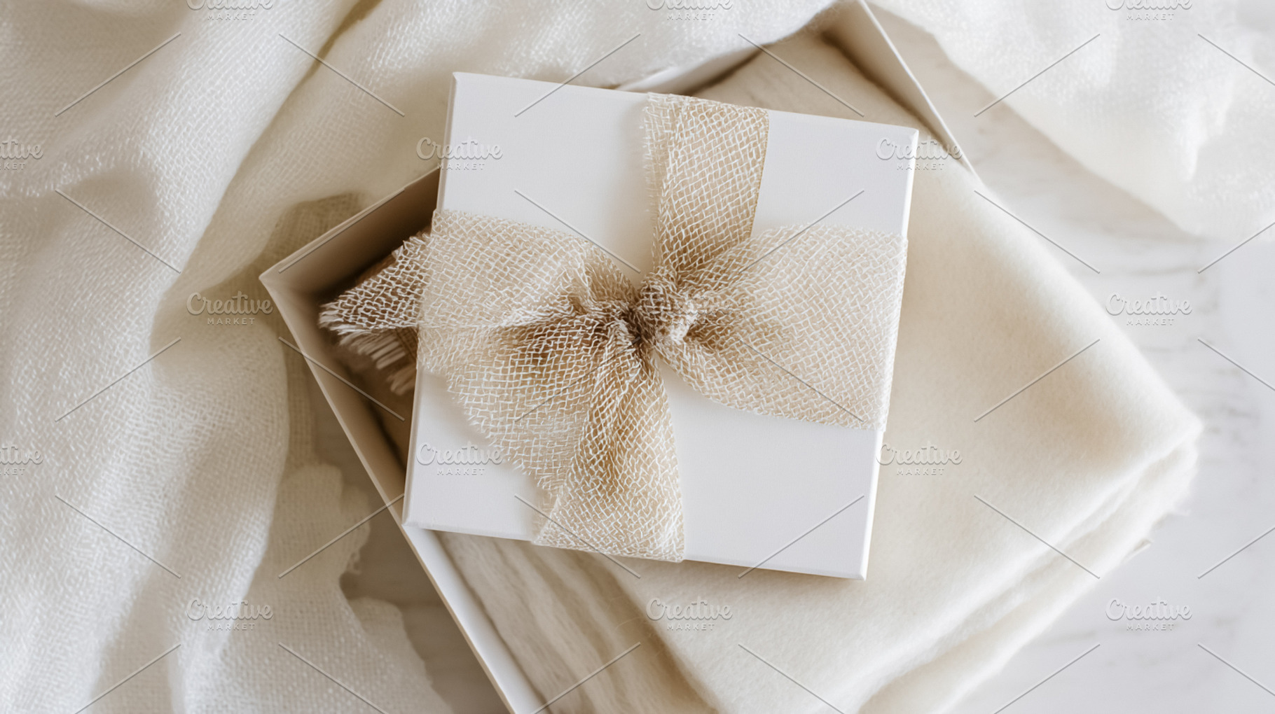 Elegant gift box with burlap ribbon bow on white textured fabric, a Photo by Studio ANNA
