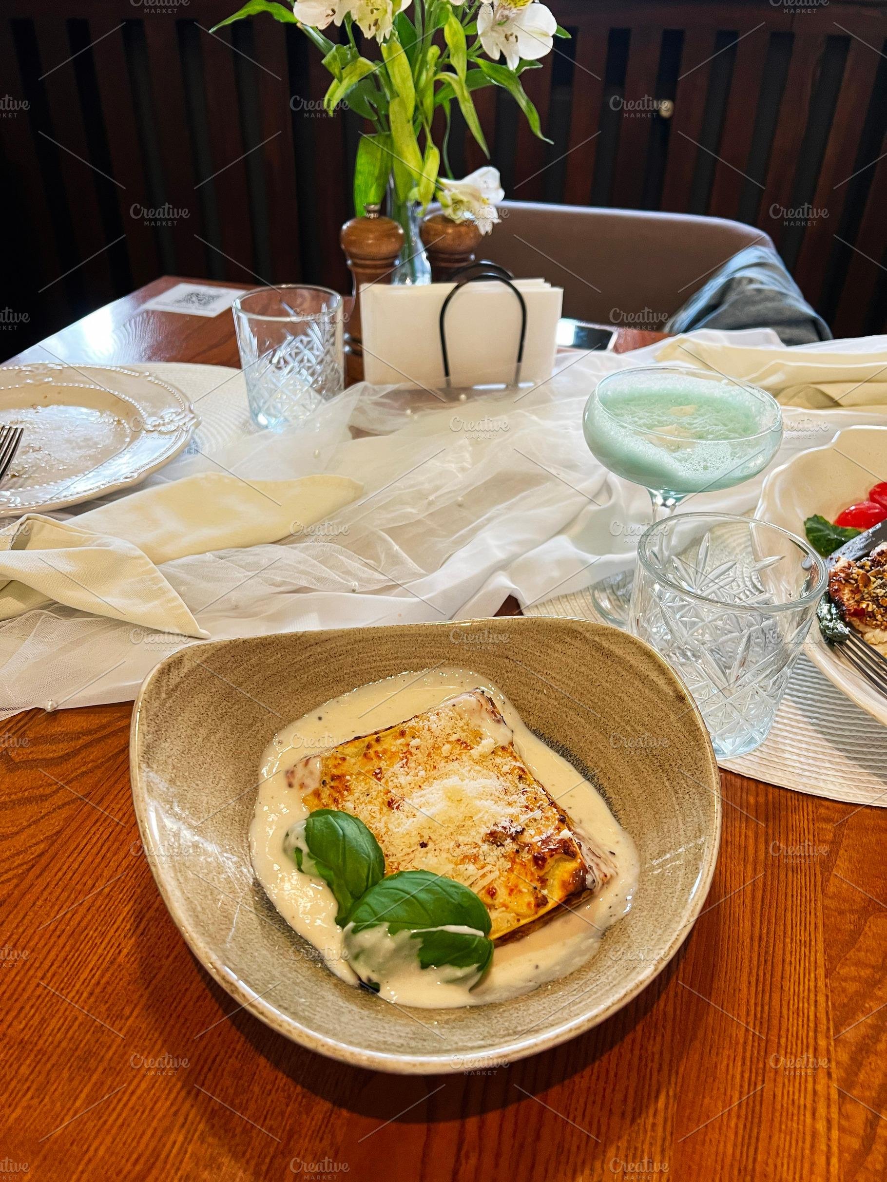 Elegant dining setup with lasagna dish, drink, and floral decoration on a table, a Photo by AS photostudio