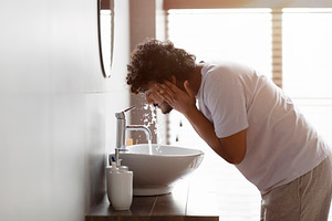 Young indian man washing face with clear water from tap bending over sink, doin, a Person Photo by Prostock-Studio