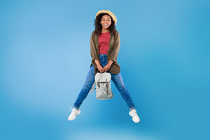 Full length portrait of positive young black woman with backpack jumping on blu, a Photo by Prostock-Studio