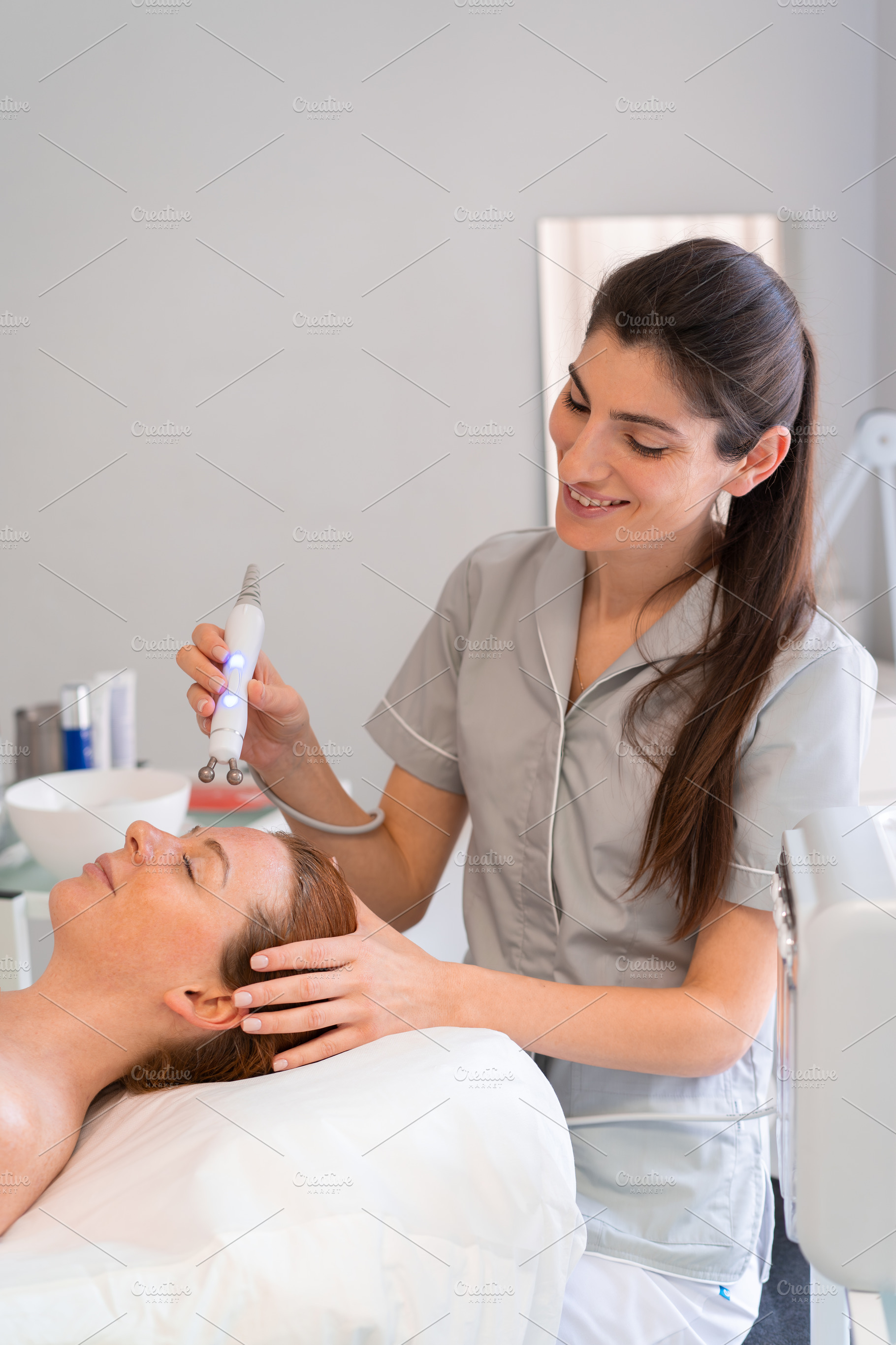 Happy cosmetologist applying iontophoresis on female client's face, a Photo by andreonegin market