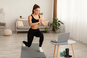 African Female Doing Forward Lunge Watching Video On Laptop Indoors, a Photo by Prostock-Studio