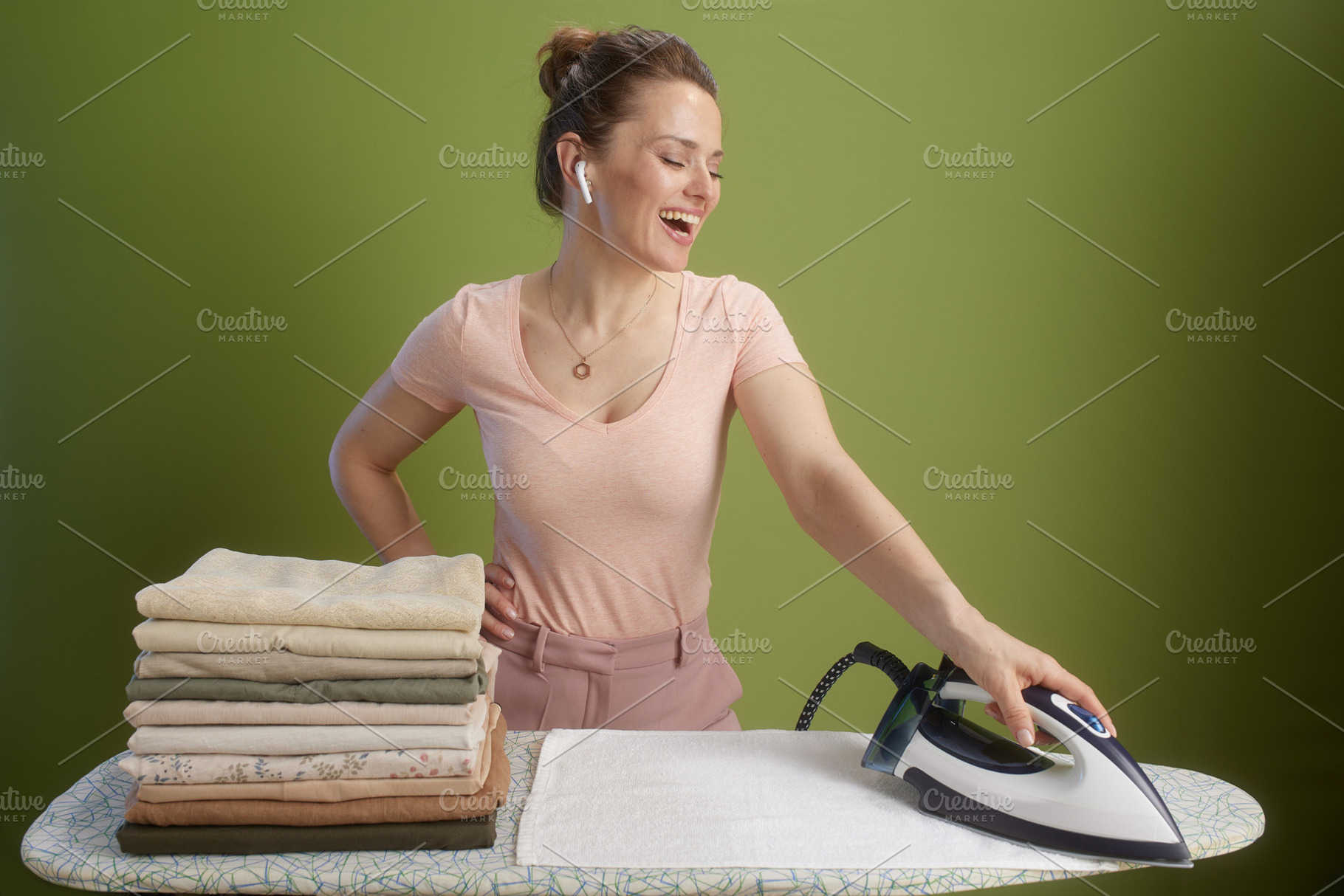 Smiling woman ironing pastel clothes, olive green backdrop, a Person Photo by Alliance Images