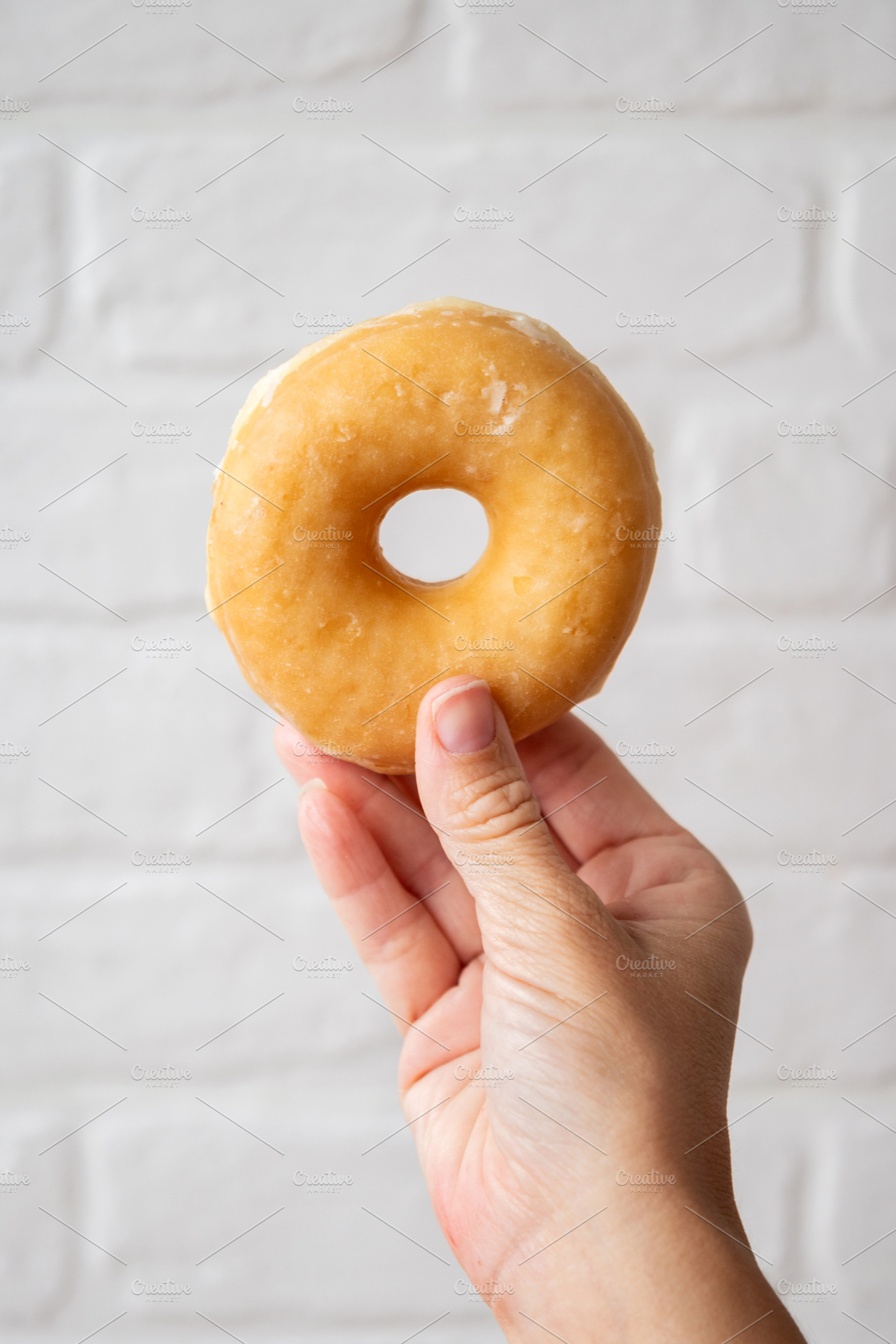 Hand Holding Glazed Donut Against White Brick Background, a Food & Drink Photo by etorres69