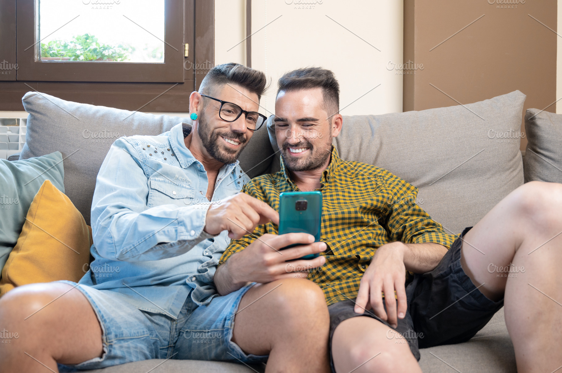Happy young gay couple using mobile phone while sitting on a sofa in the living, a Person Photo by david.herraez.calzad
