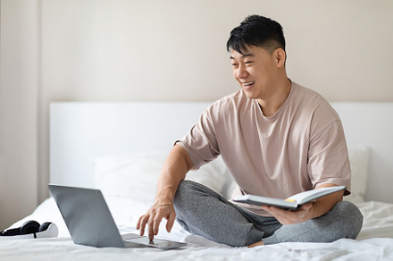 Chinese man checking notepad, using brand new laptop in bed, a Photo by Prostock-Studio