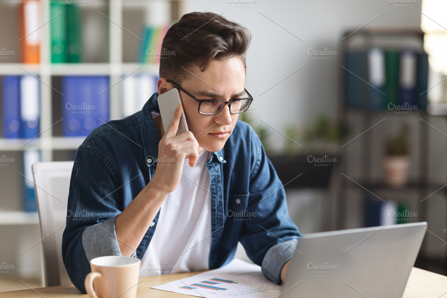 Young Man Working On Laptop And Talking On Mobile Phone In Office, a Photo by Prostock-Studio