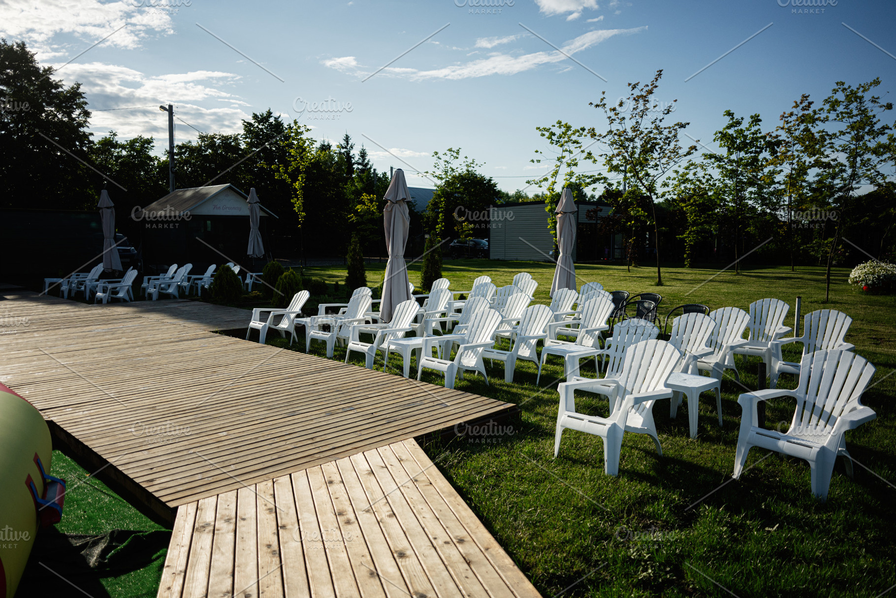 Outdoor seating area with white chairs and wooden deck in green park, a Photo by AS photostudio
