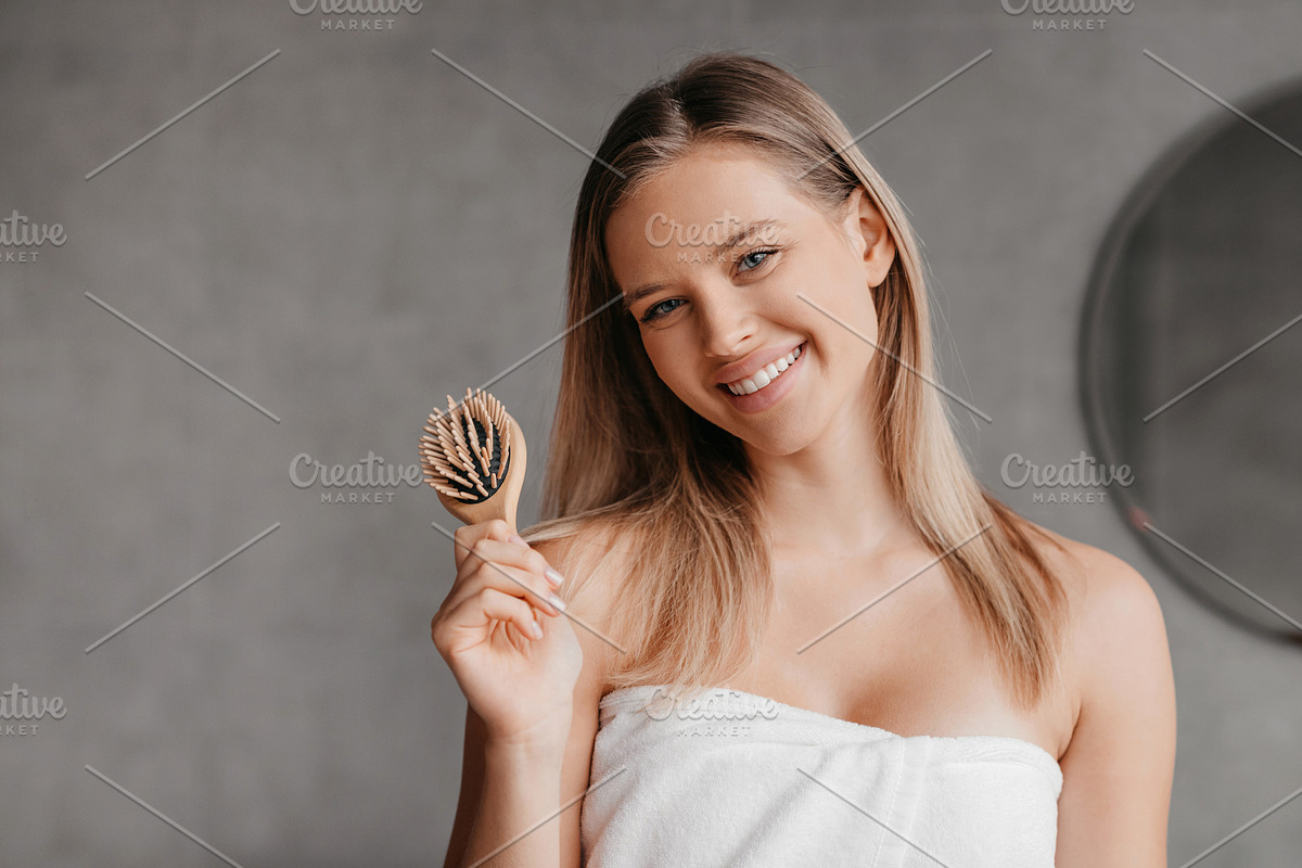 Happy young woman wrapped in white towel holding hair brush and smiling at came, a Photo by Prostock-Studio (Photo 1 of 5)