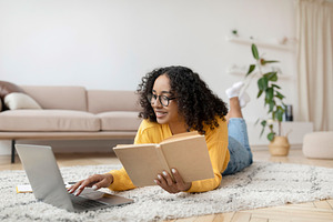 Young black woman reading book near laptop, getting ready for exam or working o, a Photo by Prostock-Studio