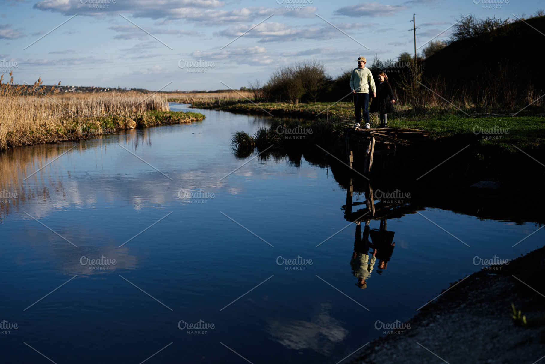 Two children standing on a small wooden dock by a calm river with reflections i, a Person Photo by AS photostudio