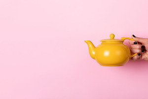 Female hand holding a yellow ceramic teapot against a pastel pin, a Food & Drink Photo by Ales Munt