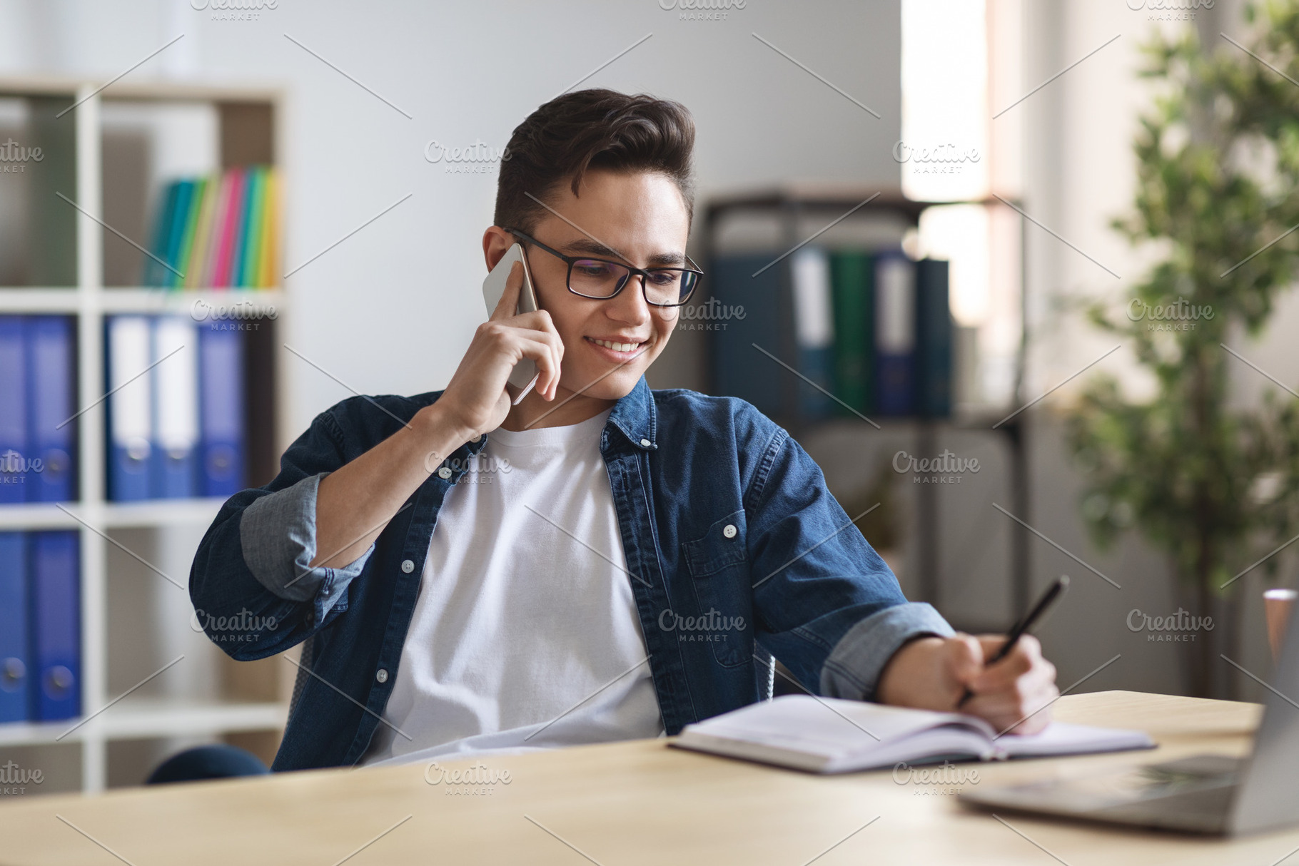 Millennial man writing notes while making phone call at workplace in office, a Photo by Prostock-Studio