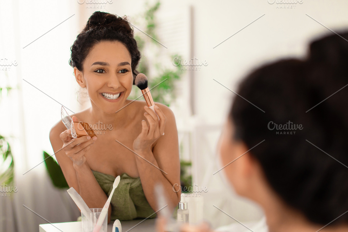 Smiling millennial woman in towel applying makeup in front of mirror, putting o, a Photo by Prostock-Studio (Photo 1 of 2)
