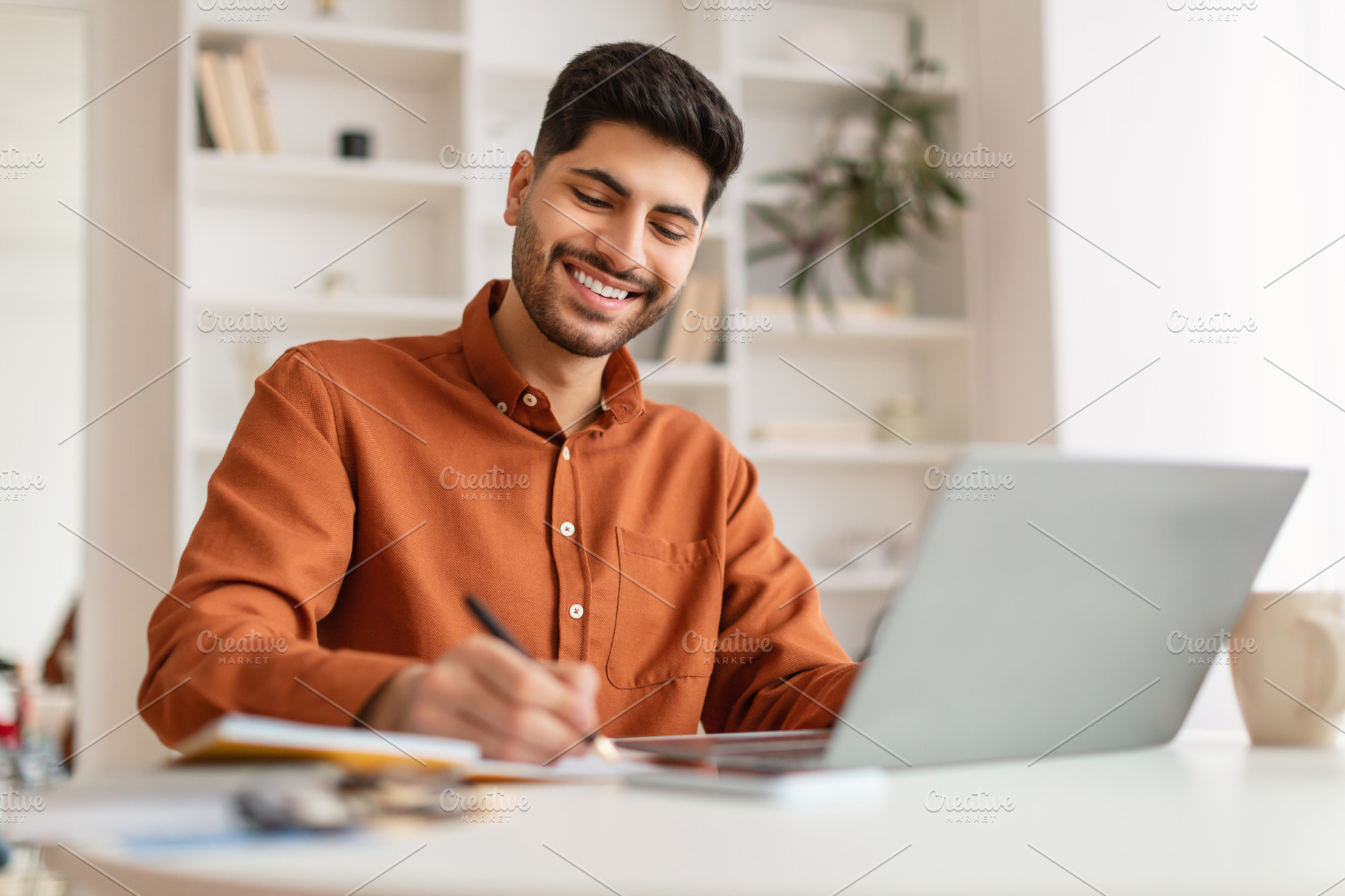 Portrait of smiling Arab man using laptop and writing, a Photo by Prostock-Studio