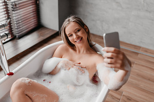 Pretty lady relaxing in foamy bath and taking selfie on smartphone while enjoyi, a Photo by Prostock-Studio