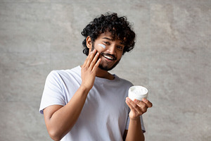 Cheerful indian man applying cream on face, holding moisturizer jar in hand, sm, a Photo by Prostock-Studio