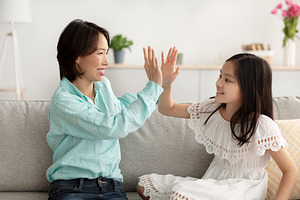 Cute little Asian girl giving high five to her grandmother, sitting on couch at, a Photo by Prostock-Studio