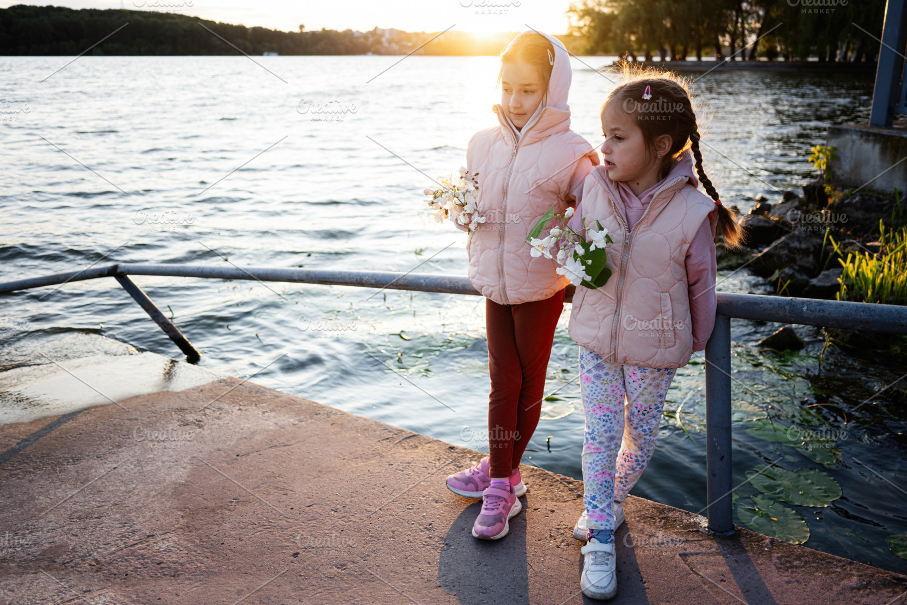 Two children holding flowers by a lakeside during sunset, a Photo by AS photostudio