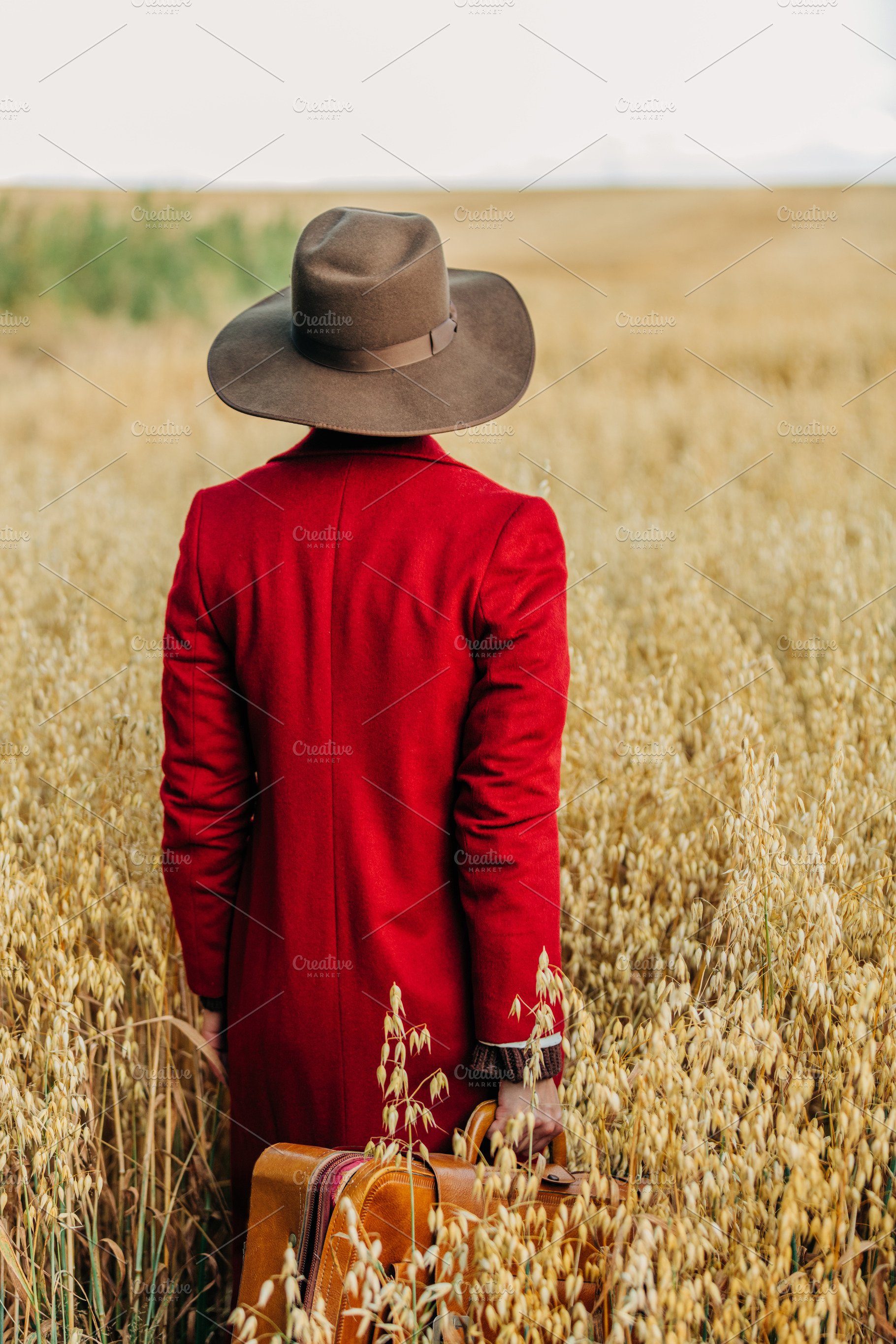 Stylish woman in vintage hat red coat with suitcase on oat field, a Person Photo by Masson Simon