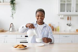 Smiling millennial african american woman in casual pouring milk at coffee at t, a Person Photo by Prostock-Studio