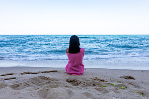 Rear view of a woman sitting alone on sandy beach facing the calm ocean under c, a Person Photo by Ales Munt