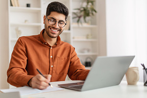 Smiling Arab man in glasses using laptop and taking notes, a Photo by Prostock-Studio