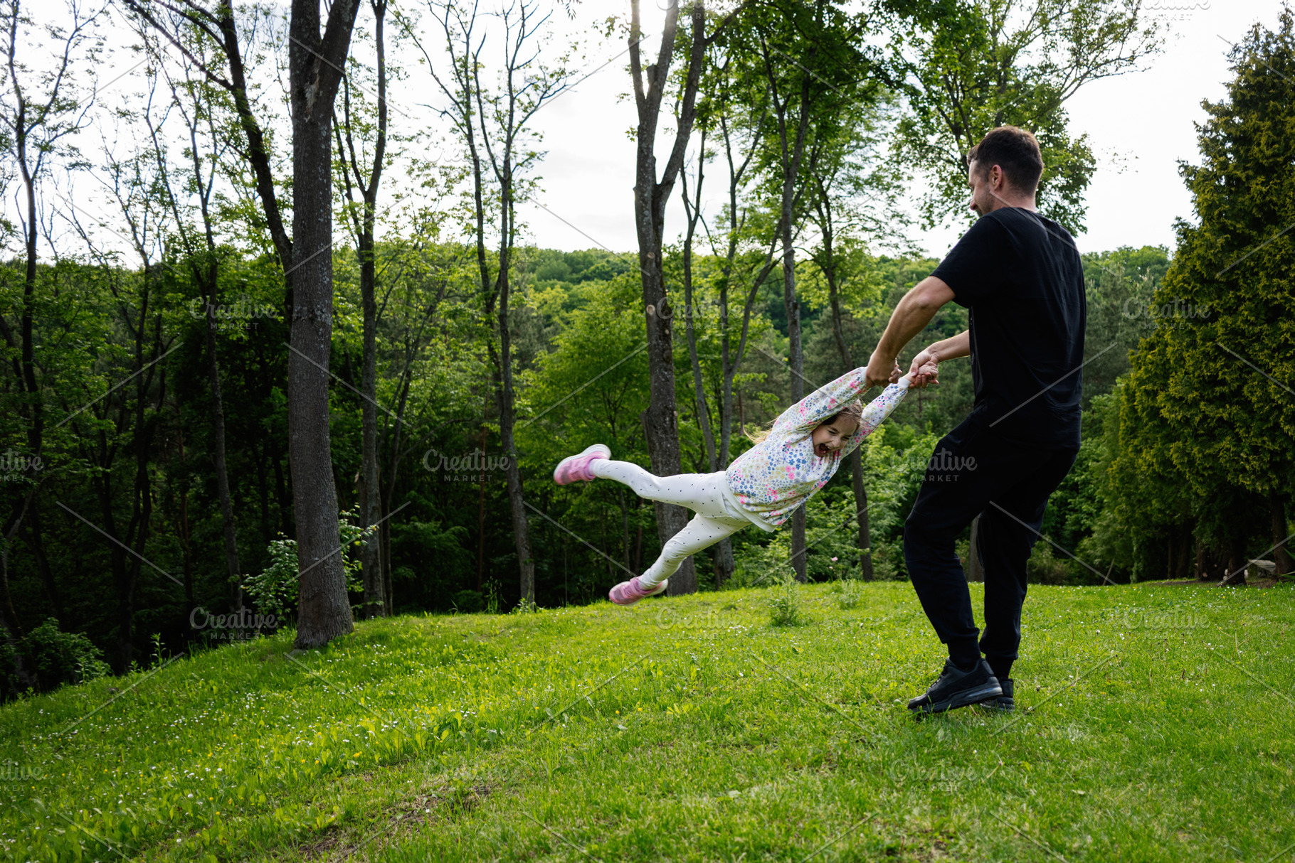 Father swinging daughter playfully in a green forested outdoor setting, a Person Photo by AS photostudio