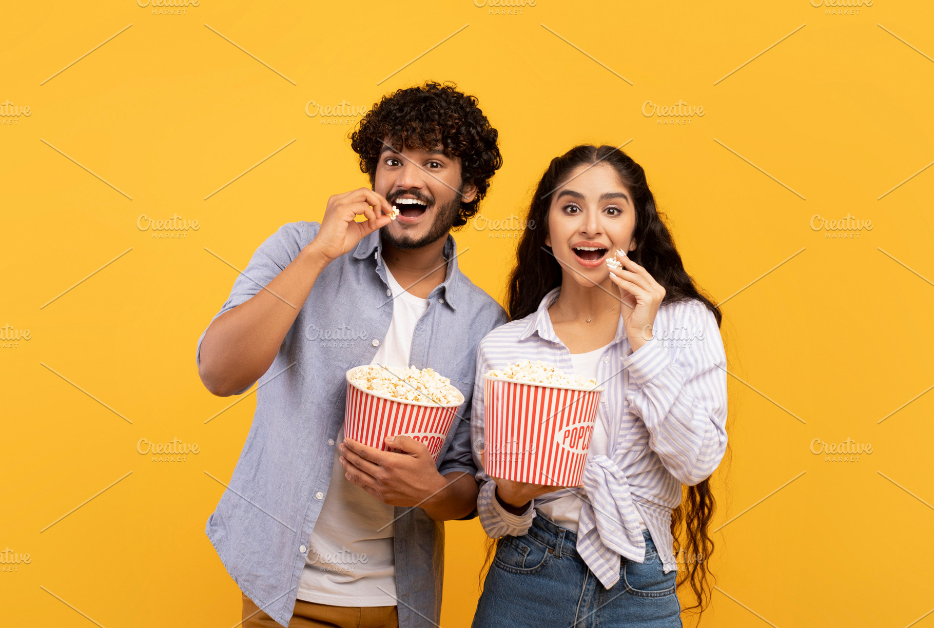 Excited indian man and woman eating popcorn at cinema, standing over yellow stu, a Person Photo by Prostock-Studio