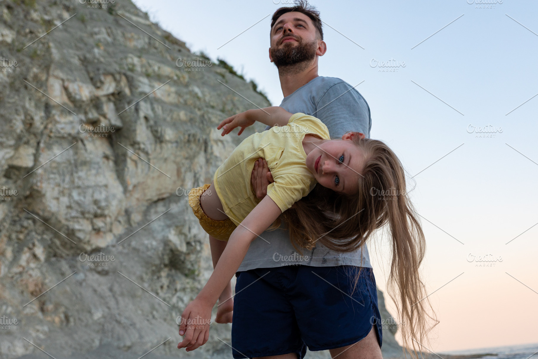 Smiling father playfully holds young daughter with long hair beside rocky cliff, a Person Photo by Ales Munt