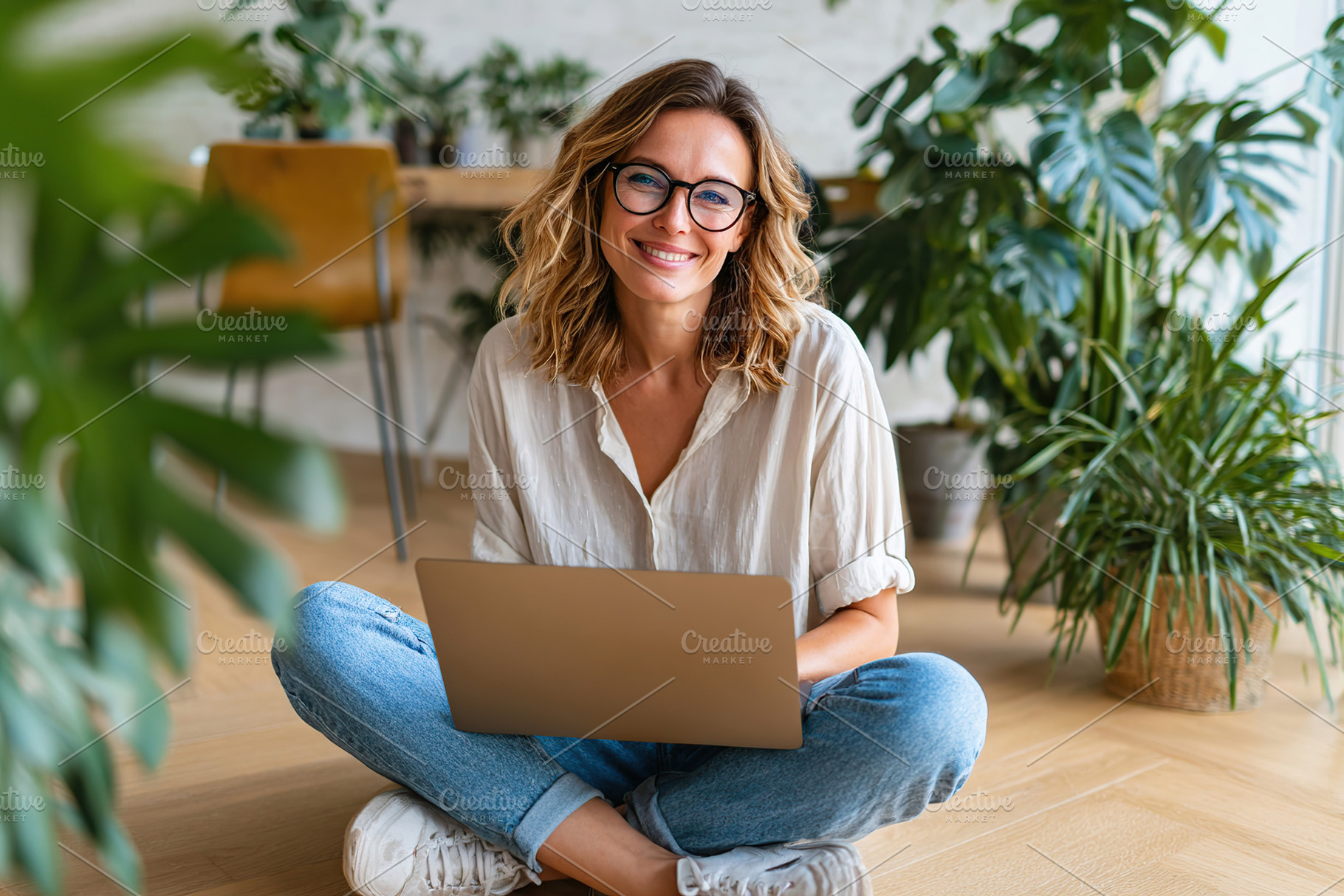 Woman sitting cross-legged on the floor using a laptop in a bright, plant-fille, a Photo by AlyaAnd