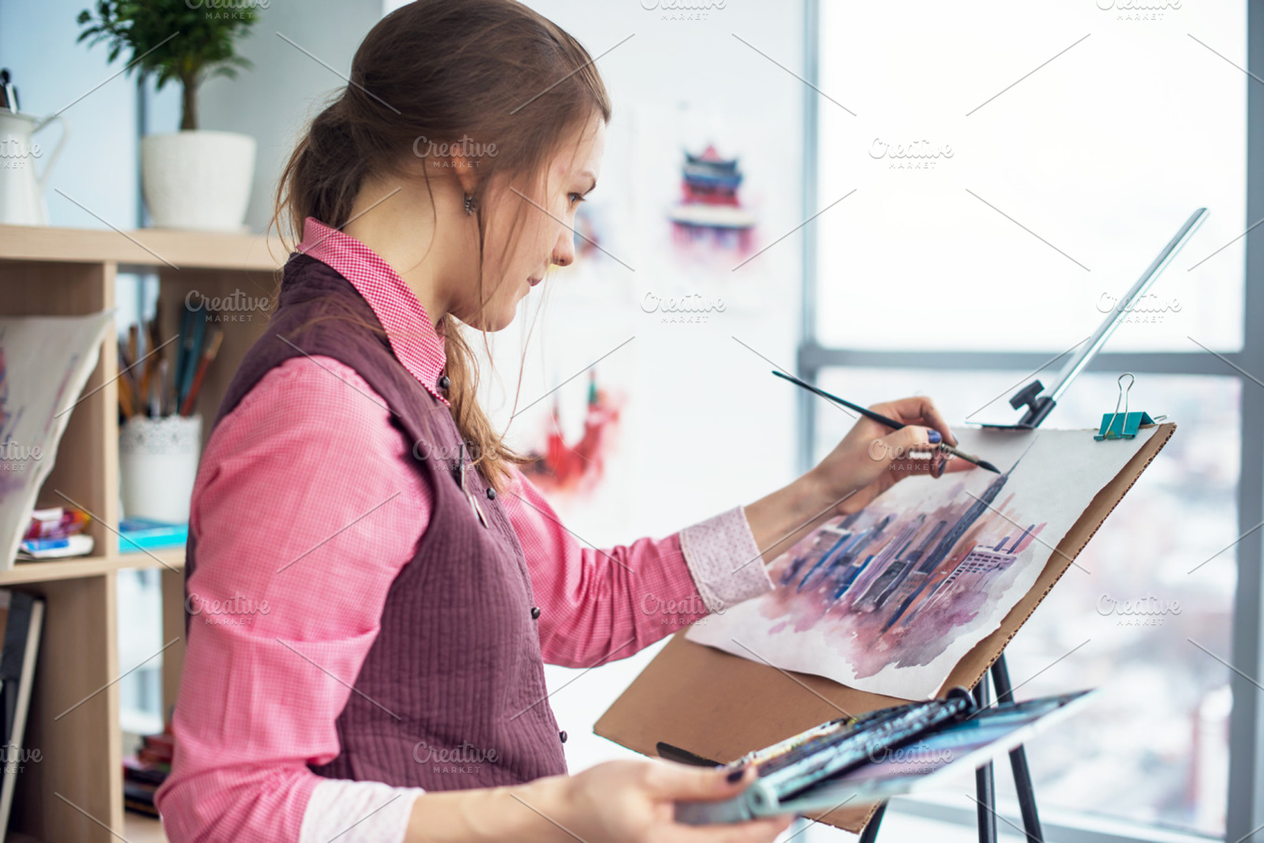 Side view portrait of a young woman painter drawing cityscape with watercolor palette on white paper using easel., an Arts & Entertainment Photo by undrey