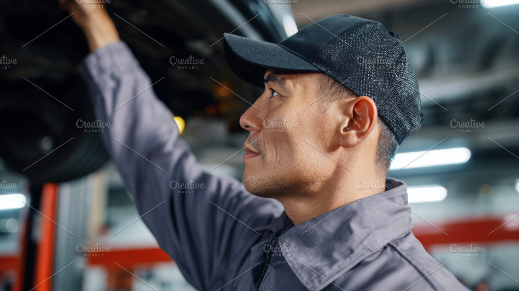 Focused Asian male mechanic inspecting a vehicle underneath in a garage, wearin, a Business Photo by ImageMir