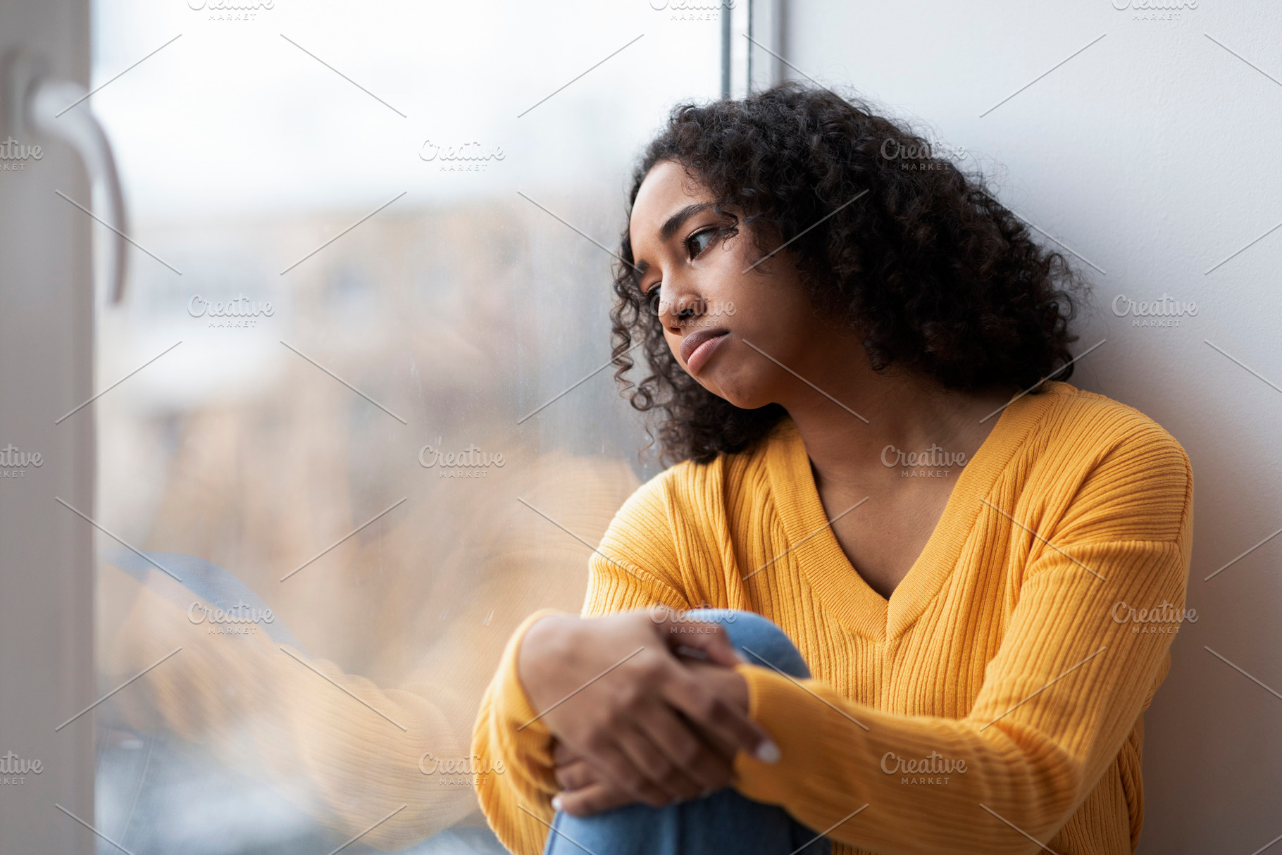 Depressed young African American woman sitting near window, looking outside on, a Photo by Prostock-Studio
