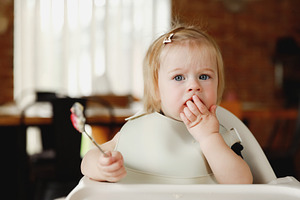 Infant girl or toddler sitting on high chair. Eating food by herself using spoo, a Person Photo by Lenor4ik