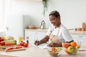 Cheerful millennial black lady in apron makes notes on table with organic veget, a Person Photo by Prostock-Studio