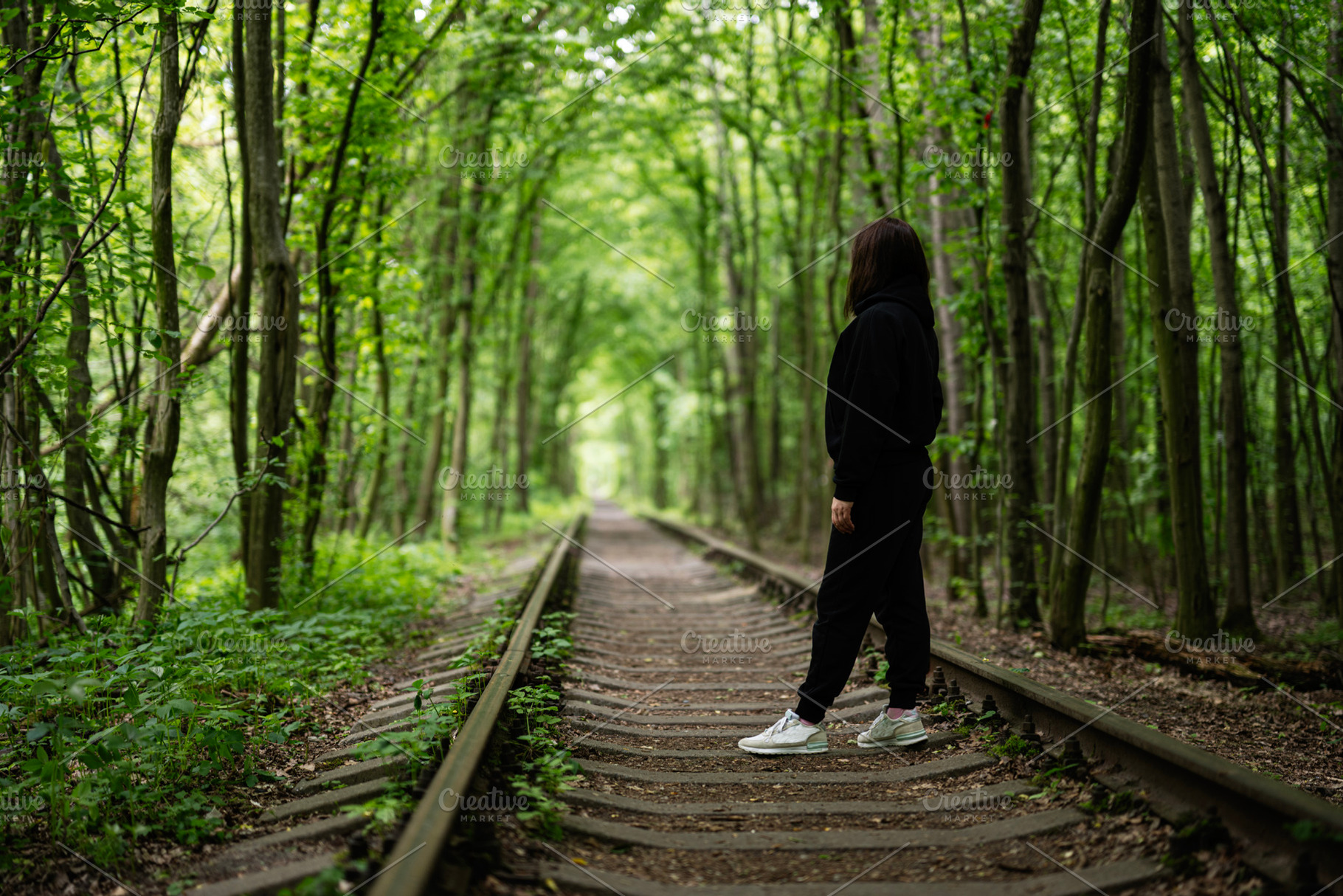 Woman walking along forest railway tracks surrounded by lush green trees, a Photo by AS photostudio