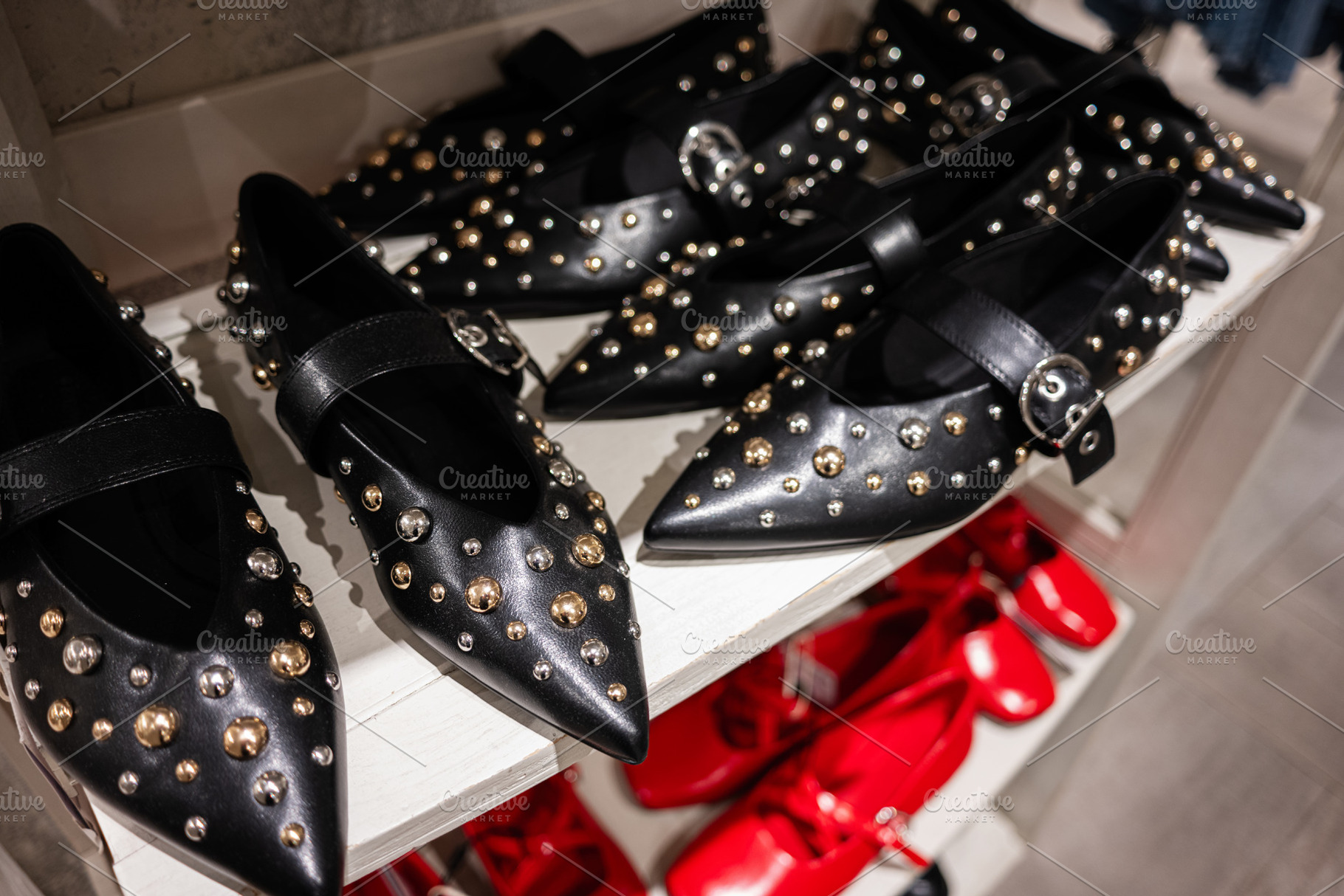 Black leather flats with studs displayed on a shelf with red shoes nearby, a Photo by AS photostudio