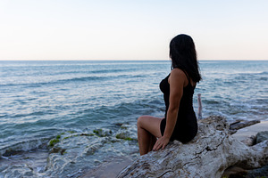 Woman in black dress sits on driftwood, gazing at the tranquil ocean under a cl, a Person Photo by Ales Munt