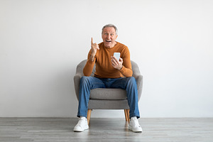 Portrait of mature man using smartphone sitting on armchair, a Photo by Prostock-Studio
