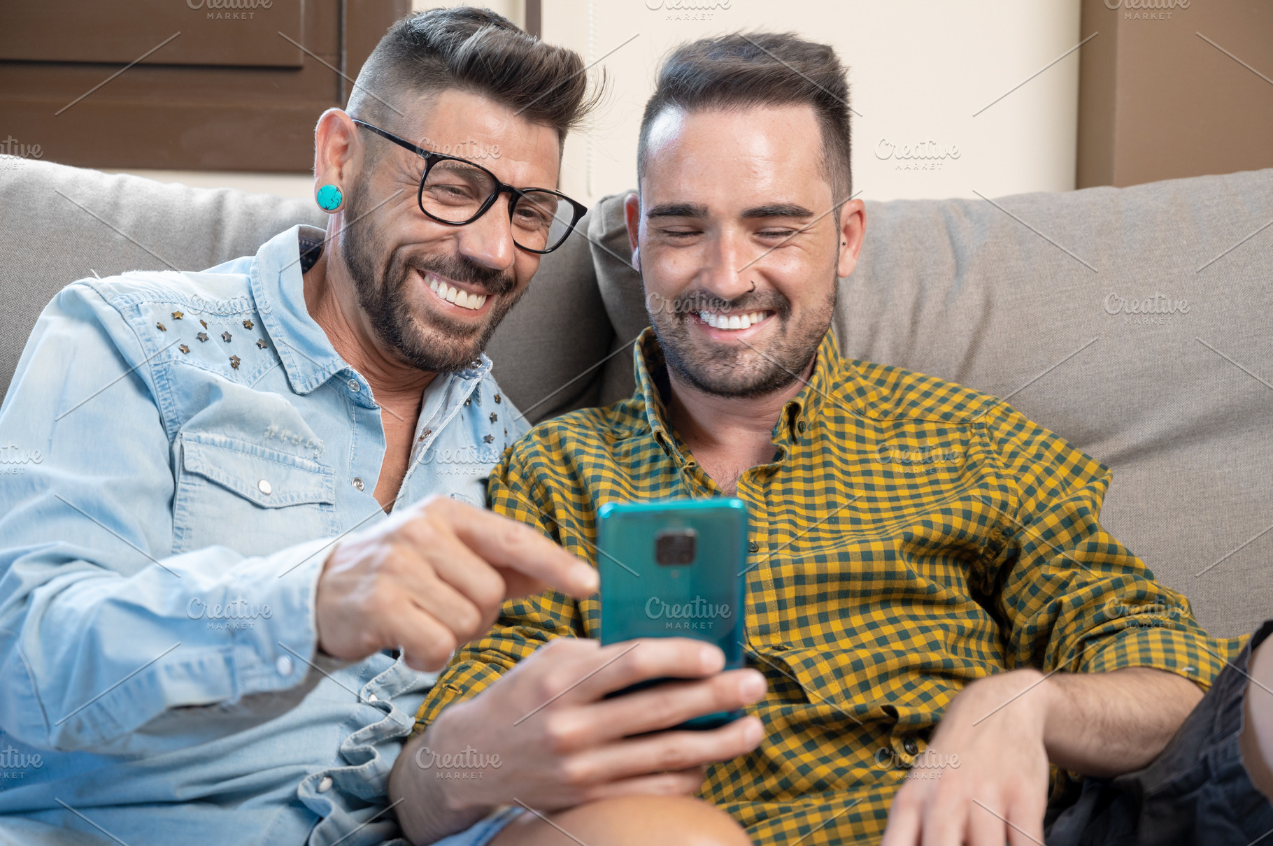 Happy young gay couple using mobile phone while sitting on a sofa in the living, a Person Photo by david.herraez.calzad