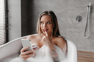 Mysterious caucasian woman using mobile phone while lying in foamy bath, textin, a Photo by Prostock-Studio