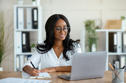 Cheerful black business lady working on laptop, taking notes during online work, a Photo by Prostock-Studio