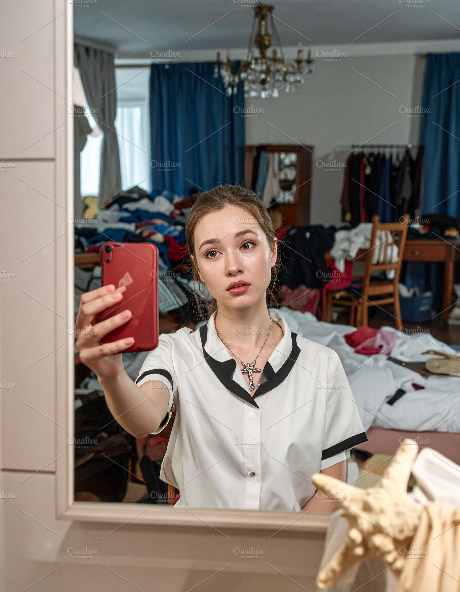Young woman holding a phone takes a mirror selfie in a cluttered, a Person Photo by DanFLCreativo