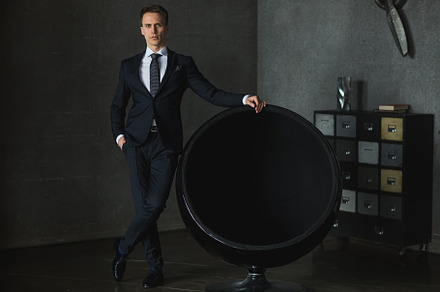 Young businessman in black suit posing near the chair on gray background