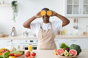 Glad funny millennial black female in apron have fun, puts orange slices to eye, a Person Photo by Prostock-Studio