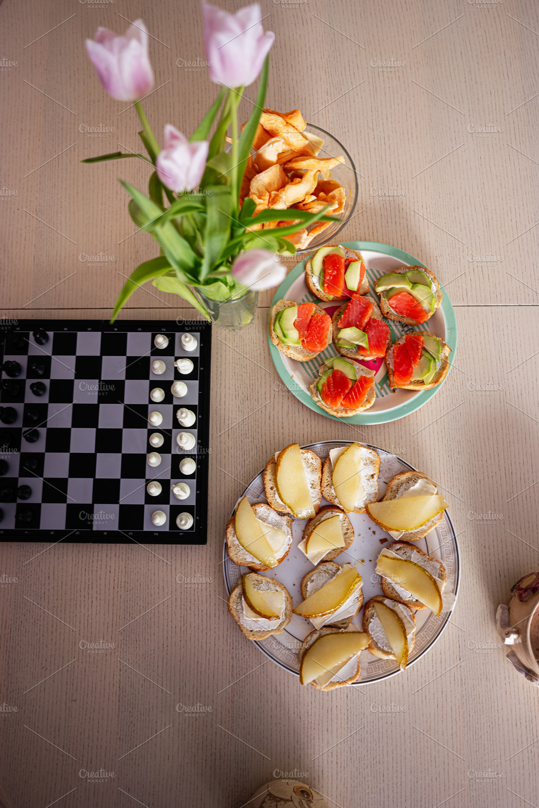 Assorted snacks and chess on a wooden table next to fresh tulips, a Photo by AS photostudio