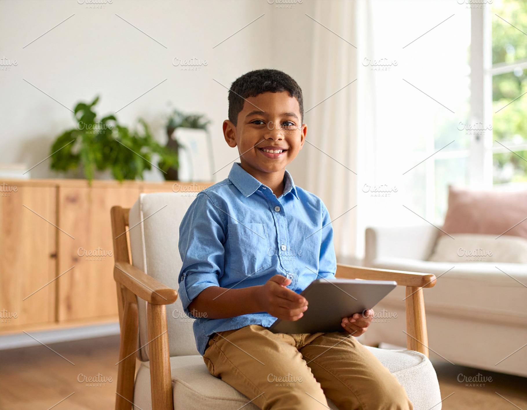 Boy with a tablet sitting on chair at living room while smiling, a Person Photo by DanFLCreativo