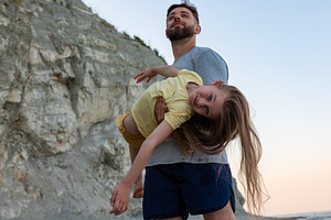 Smiling father playfully holds young daughter with long hair beside rocky cliff, a Person Photo by Ales Munt
