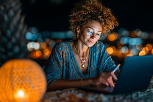 Woman working on laptop outdoors with warm lantern lighting at nighttime, a Business Photo by AS photostudio